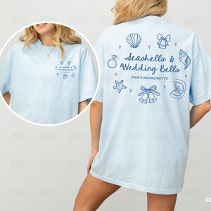 K&ouml;nnte beinhalten: Hellblaues T-Shirt mit dem Text "Seashells & Wedding Bells" und zugeh&ouml;rigen Grafiken. Das Shirt hat auch den Text "Julie's Bachelorette" und ist Gr&ouml;&szlig;e L.