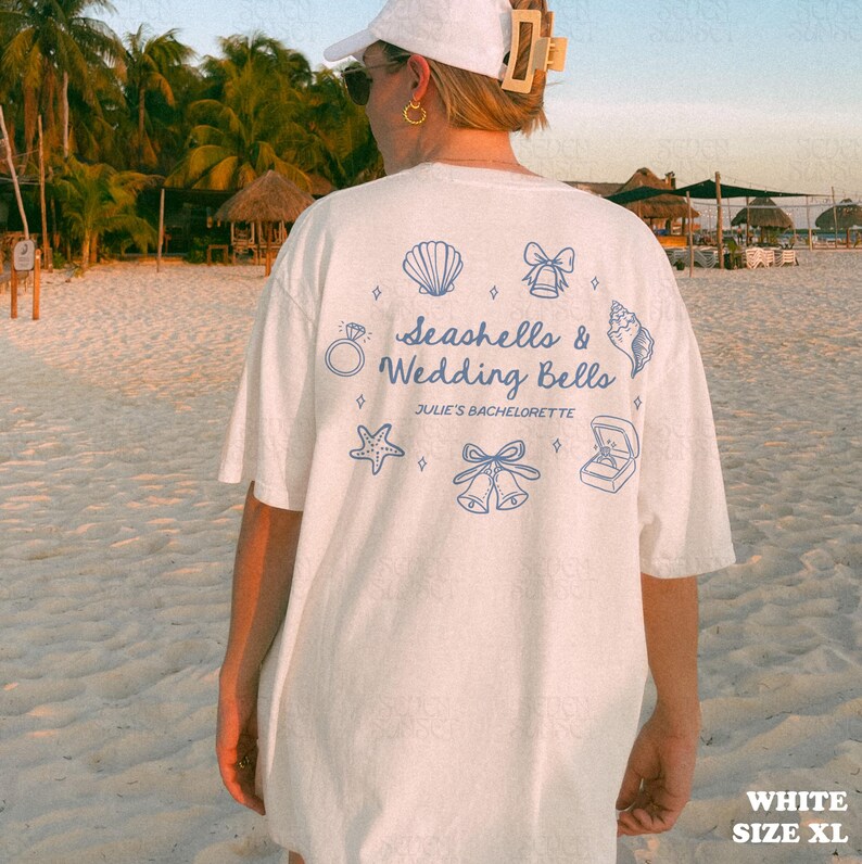 K&ouml;nnte beinhalten: Wei&szlig;es T-Shirt mit blauen Muschel- und Hochzeitsmotiven. Der Text "Seashells & Wedding Bells" und "Julie's Bachelorette" ist auf der R&uuml;ckseite aufgedruckt. Das Shirt ist in Gr&ouml;&szlig;e XL.