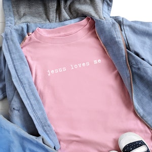 Op de afbeelding: Een roze longsleeve t-shirt met de tekst "jesus loves me" in witte letters.