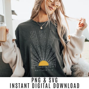 Puede incluir: Una camiseta gris oscuro con un diseño de rayos de sol blancos y el texto "Sé la luz Mateo 5:14".