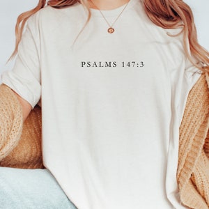 以下が含まれることがあります： 前面に「PSALMS 147:3」という文字がプリントされた白いTシャツです。