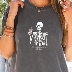 Puede incluir: Camiseta gris con un esqueleto blanco sosteniendo una taza de café. El texto "Dry bones come alive Ezekiel 37" está impreso debajo del esqueleto.