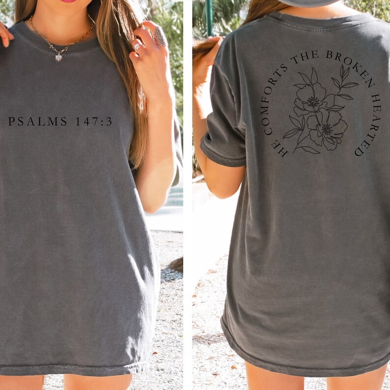 以下が含まれることがあります： 前面に「PSALMS 147:3」の文字、背面に花柄のデザインと「HE COMFORTS THE BROKEN HEARTED」の文字が書かれたグレーのTシャツです。