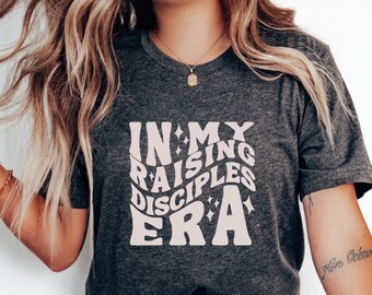 May include: A dark gray t-shirt with a white graphic that says "In My Raising Disciples Era" in a retro font with stars.