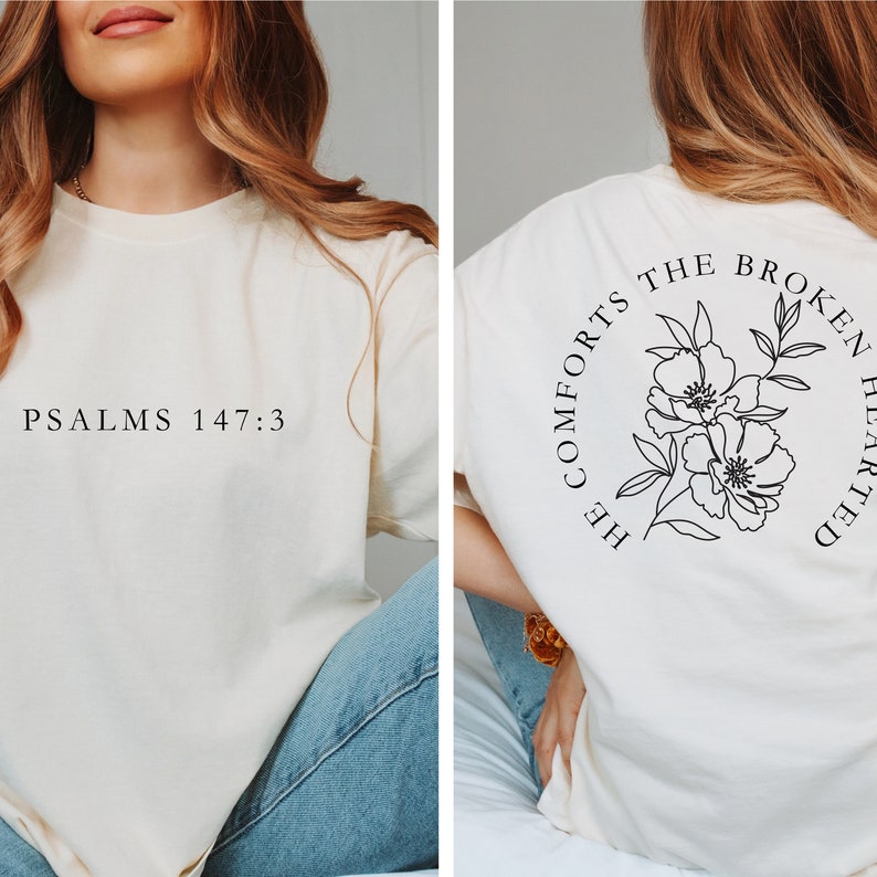 以下が含まれることがあります： 前面に「PSALMS 147:3」の文字、背面に「HE COMFORTS THE BROKEN HEARTED」の文字と花柄のデザインが施された白いTシャツです。