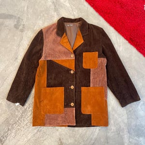 Puede incluir: Chaqueta de pana patchwork en tonos marrón, óxido y beige. La chaqueta tiene cuello, cierre de botones y dos bolsillos delanteros. La chaqueta tiene una estética vintage.