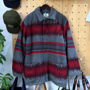 Può includere: Giacca di lana grigia con motivo geometrico rosso e grigio. La giacca ha una chiusura con bottoni, due tasche e un colletto. È appesa a una gruccia di legno contro una pannello forato di legno.