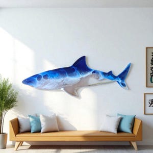 Op de afbeelding: Een grote, aan de muur gemonteerde haai sculptuur met een opvallend blauw en wit ontwerp, dat op oceaangolven lijkt. Het lichaam van de haai is een mix van doorschijnende en ondoorzichtige materialen, wat een dynamisch visueel effect creëert.