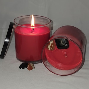 May include: Two red candles in clear glass jars. One candle is lit, with a flame burning. The other candle is tipped over, revealing a black stone and a brown and white stone. A black lid is next to the lit candle.