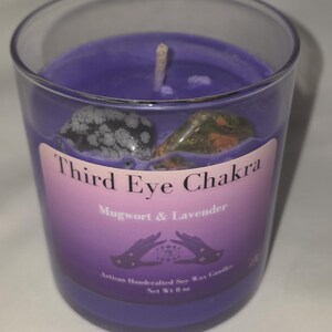 Puede incluir: Una vela de cera de soja morada en un vaso transparente, etiquetada como "Third Eye Chakra" con el texto "Mugwort & Lavender". La vela contiene dos piedras y una mecha. La etiqueta también incluye el texto "Artisan Handcrafted Soy Wax Candles" y "Peso neto 227 g."