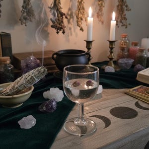 May include: A mystical scene featuring a table with a glass of water, tarot cards, and an open book. Lit candles, crystals, and dried herbs create a spiritual ambiance. A sage bundle sits in a bowl, with smoke rising.
