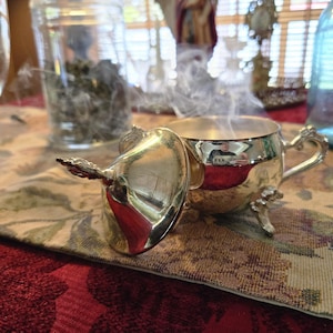 May include: A polished silver-toned metal sugar bowl with a matching lid resting beside it. The bowl features a rounded body, a single curved handle, and three ornate feet. Wisps of white smoke rise from the open top of the container.