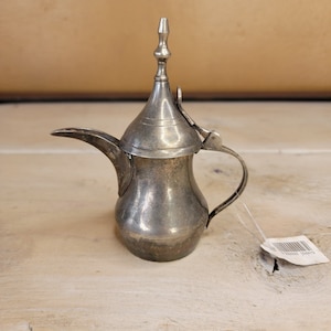 May include: A silver-colored metal teapot with a decorative spout and a hinged lid. The teapot has a traditional Arabic design with a tall, pointed lid.