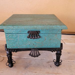 Op de afbeelding: Een teal houten doos met een decoratief metalen scharnier en een zwarte metalen standaard met vier poten. De doos staat op een licht houten oppervlak.