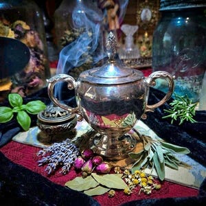 May include: A silver-coloured, ornate lidded container with handles, surrounded by herbs, flowers, and a small incense burner. Wisps of smoke rise from the container, suggesting use. The scene is set on a patterned surface with a dark, velvety cloth.