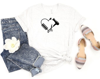 May include: A white t-shirt with a black heart design featuring a hair dryer, comb, and scissors. The shirt is displayed on a white background with a pair of blue jeans and a pair of patterned shoes.