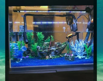 May include: An aquarium with a sunken ship, artificial plants, and blue gravel. The tank features a variety of decorations, including a treasure chest, coral, and a background structure. The water is clear, and the tank is illuminated by a light.