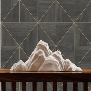 May include: White ceramic mountain sculpture on a dark wood table with a geometric pattern wallpaper in the background. The wallpaper has a dark grey background with gold lines forming triangles.