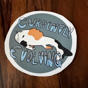 May include: A round sticker with a cartoon illustration of a white, black, and orange animal. The text "CURRENTLY EVOLVING" is written in blue, bubble-style letters around the animal on a gray background.