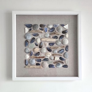 May include: A square white frame containing a collection of various seashells, pieces of driftwood, and small fragments of sea glass arranged on a light beige linen background. The shells are in shades of grey, white, blue, and brown.