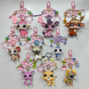 May include: A selection of animal-themed keyrings. Each keyring features a small plastic animal figure, including a cat, cow, and cheetah, with decorative pink bows and star-shaped details. The keyrings are attached to a metal clasp.