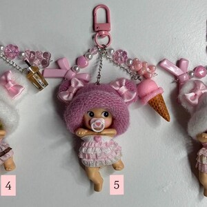 May include: Three pink keychains with baby doll figures wearing pink outfits and hats, each with a dummy. The keychains have pink clips, decorative beads, and miniature accessories such as a drink or ice cream cone.