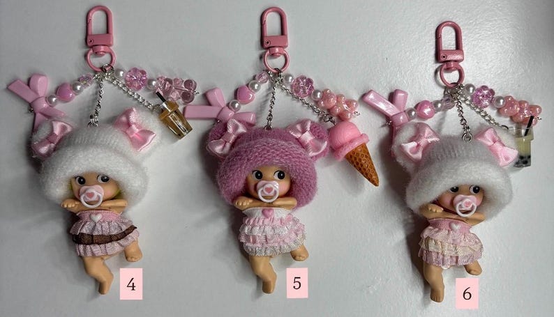 May include: Three pink keychains with baby doll figures wearing pink outfits and hats, each with a dummy. The keychains have pink clips, decorative beads, and miniature accessories such as a drink or ice cream cone.