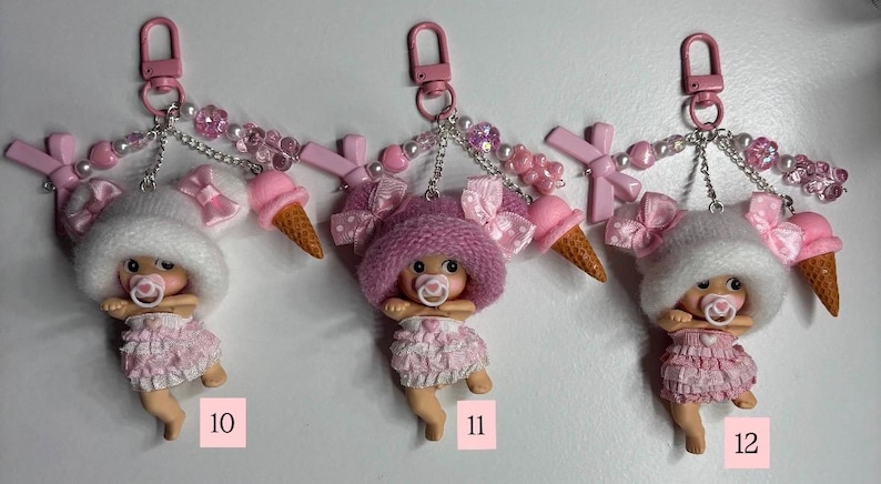 May include: Three keyrings with baby doll charms. Each doll wears a pink dress and hat, and has a dummy. The keyrings have pink clasps, decorative beads, bows, and a miniature ice cream cone. Numbers 10, 11, and 12 are visible.