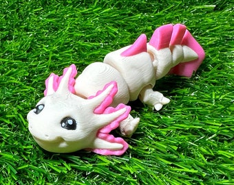 May include: A white and pink axolotl figurine with black eyes and pink gills. The figurine is lying on a bed of green artificial grass. The axolotl has a detailed, textured design.