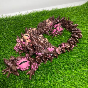 May include: A brown, segmented creature with multiple pink and black eyes and a series of appendages. The creature is positioned on a bed of green grass. The eyes have a pink iris and black pupil.