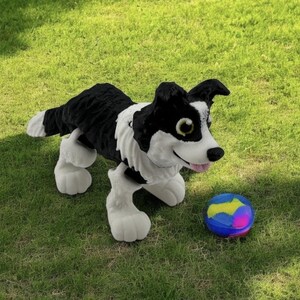 May include: A plush black and white dog toy with a smiling face and a pink tongue, positioned on green grass next to a colorful ball. The dog has large, expressive eyes and white paws.