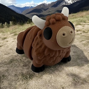 May include: A brown, cartoon-style bull sculpture with white horns and black hooves. The bull has a smiling face and is set against a backdrop of mountains and a grassy field. The sculpture appears to be made of layered wood.