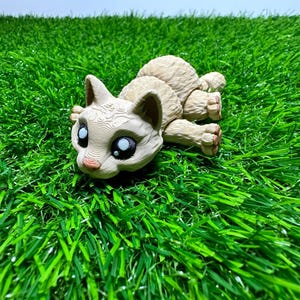 May include: A beige cat figurine with large blue eyes and a pink nose, lying on a bed of green artificial grass. The cat's fur has a detailed, textured design. The figurine is a decorative item.