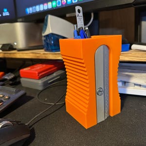 May include: Orange plastic pencil holder with a built-in pencil sharpener. The sharpener has a silver blade and a silver screw. The holder has a ribbed design.