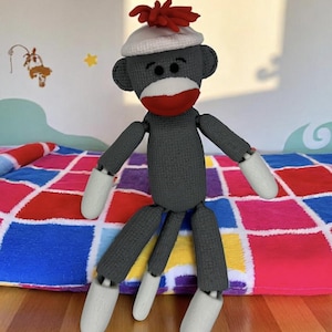 May include: A handmade sock monkey doll with a gray body, white hands and feet, and a red mouth. It wears a white hat with a red pom-pom. The doll is sitting on a colorful patchwork quilt.