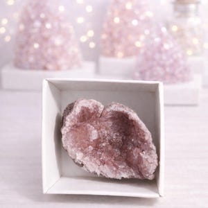 Könnte beinhalten: Eine rosa Amethyst-Geode in einer weißen Box. Die Geode hat eine raue, strukturierte Oberfläche und ein kristallines Inneres. Im Hintergrund befinden sich weitere rosa Kristallformationen und weiche Lichter.