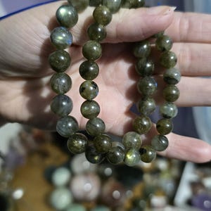 May include: A pair of Labradorite beaded bracelets. The beads are a mix of olive green and gray with iridescent flashes. The bracelets are strung on a blue cord. The background is blurred, but appears to be a collection of other gemstones.