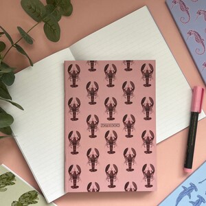 May include: A pink notebook with a lobster pattern and the word "NOTEBOOK" printed on the front cover.
