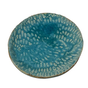 May include: A round ceramic plate featuring a vibrant turquoise glaze with a textured surface pattern of raised, elongated, teardrop-shaped indentations. The edges of the plate are slightly irregular and finished with a thin, brownish-gold rim.