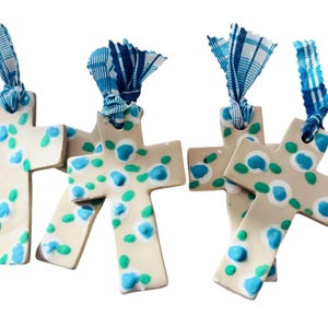 Puede incluir: Cuatro cruces de cerámica con base crema, adornadas con patrones florales azules y verdes. Cada cruz está atada con una cinta a cuadros azul y blanca. Las cruces están dispuestas sobre un fondo blanco.