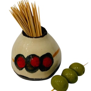 May include: A ceramic toothpick holder with a cream-colored base and a black rim. The holder features three red circles with black outlines. A bundle of wooden toothpicks is inside. Three green olives are skewered on a toothpick.