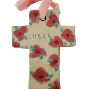 May include: A ceramic cross ornament with a cream background, adorned with red floral designs and teal accents. The word "NELL" is inscribed on the cross. A pink ribbon is attached for hanging.