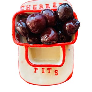 May include: A ceramic cherry pit bowl with a red trim and the word "CHERRIES" on top. The bowl is filled with dark red cherries, and the word "PITS" is written on the front.