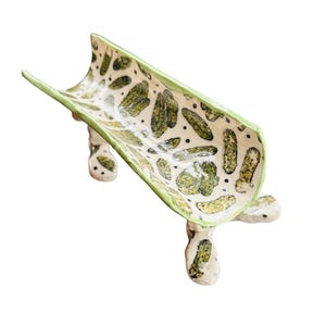 May include: A ceramic serving dish with a unique design. The dish is white with a green rim and features a pattern of green pickle illustrations. It is supported by three small, rounded legs.