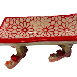 May include: A whimsical, red and white ceramic bench with a floral pattern. The bench is supported by two legs wearing pink sandals with red straps. A decorative piece for home decor.