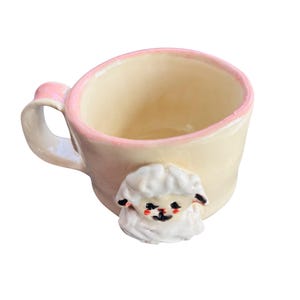 May include: Cream-colored ceramic mug with a pink rim and handle. A white sheep figurine is attached to the side of the mug. The sheep has black eyes and a black nose. The mug is designed for beverages.