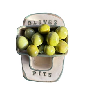 Può includere: Un piatto per olive in ceramica con le parole "OLIVES" e "PITS" in lettere nere. Il piatto è pieno di olive verdi. Il piatto è di colore beige chiaro con un bordo verde.