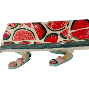 May include: A whimsical bench figurine with a watermelon pattern on the seat. The bench has legs and feet wearing pink and blue sandals. The bench is painted with a green and white color scheme.