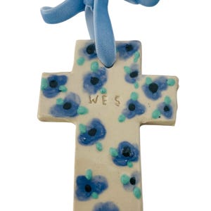 May include: A white ceramic cross with blue and green floral designs. The cross has the name "WES" inscribed on it and is attached to a blue velvet ribbon.