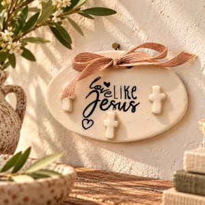 May include: An oval-shaped cream-colored wall plaque featuring the black text "love LIKE Jesus" with a small heart icon. The plaque is decorated with three small raised white crosses and a tied tan burlap ribbon at the top center.